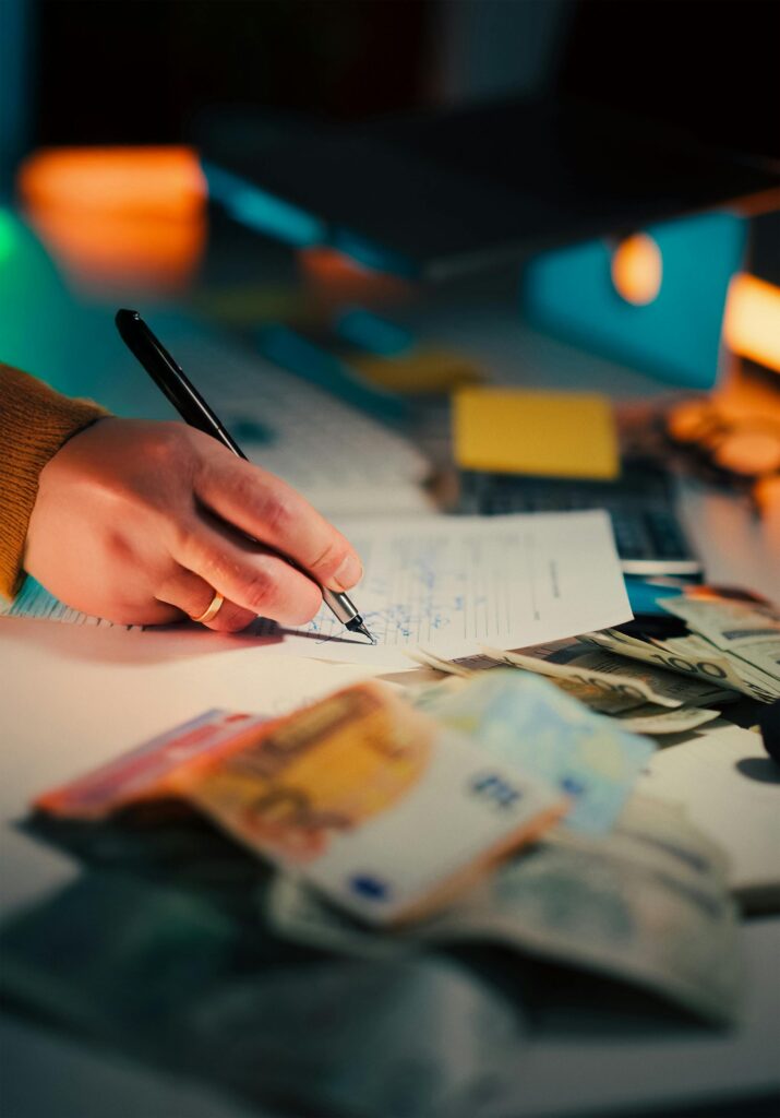 Credito y Finanzas Close-up of a hand writing on documents with euro notes scattered on a desk, focusing on finance and business.